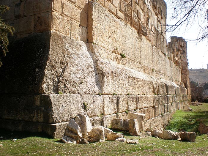 The podium wall of the Temple of Jupiter at Baalbek seen at a sharp angle along its length: an immense single course of pale limestone runs unbroken into the distance, dwarfing the regular smaller blocks stacked above and below it, with grass and fallen rubble at its base