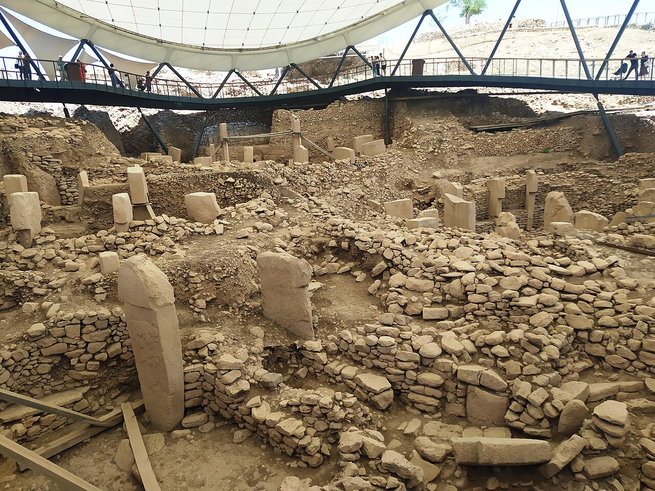 Excavated circular stone enclosures at Göbekli Tepe seen from a raised walkway: rings of upright T-shaped limestone pillars set into low drystone walls, with visitors walking on a steel bridge overhead beneath a white tensioned fabric shelter roof