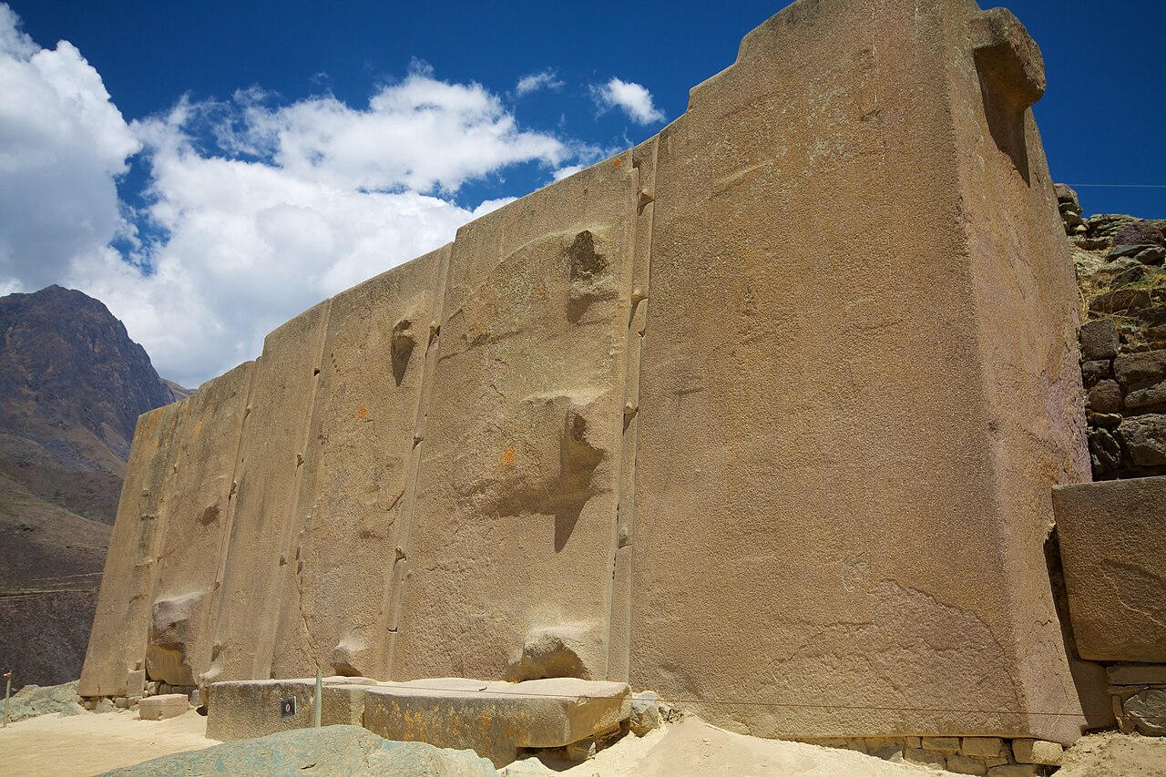 A row of six enormous upright stone slabs forming a wall at Ollantaytambo, their pale surfaces smoothed but interrupted by several rough protruding lumps, standing against a deep blue sky with white clouds and a mountain ridge behind