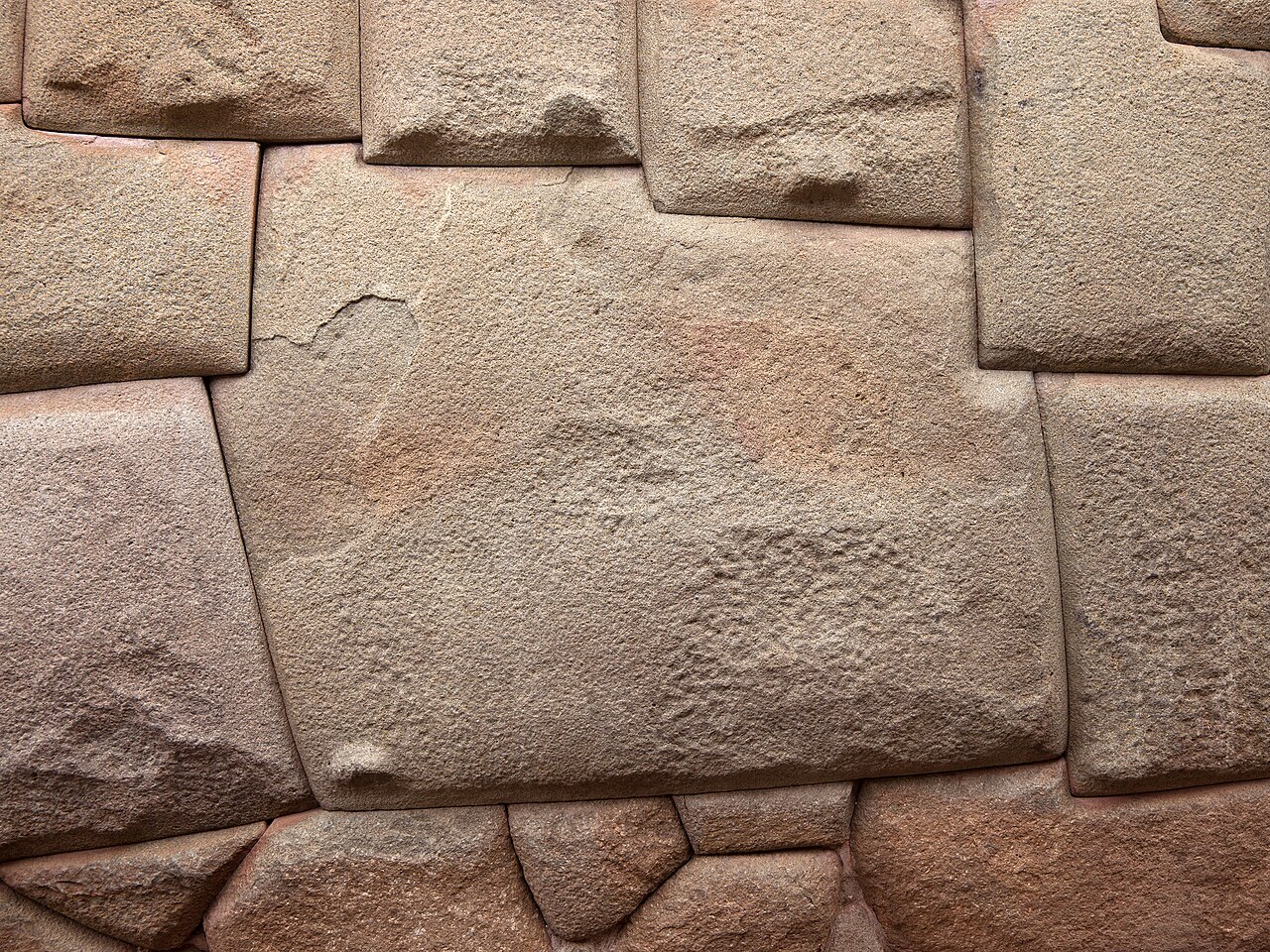 Close view of an Inca wall in Cusco: a large pale stone with twelve distinct angled sides fitted exactly among surrounding blocks, every joint a fine dark line, the stone faces gently pillowed and rounded