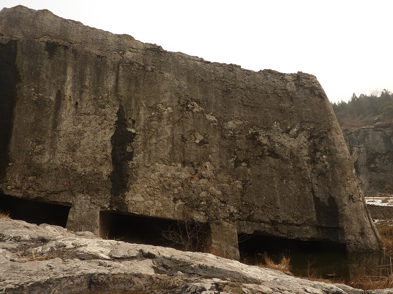 The unfinished stele base at Yangshan Quarry: a vast rectangular mass of grey worked stone, its front face cut smooth and vertical, undercut along the bottom edge where several square pillars of uncut rock have been left standing to hold it up, with rough quarry floor in the foreground
