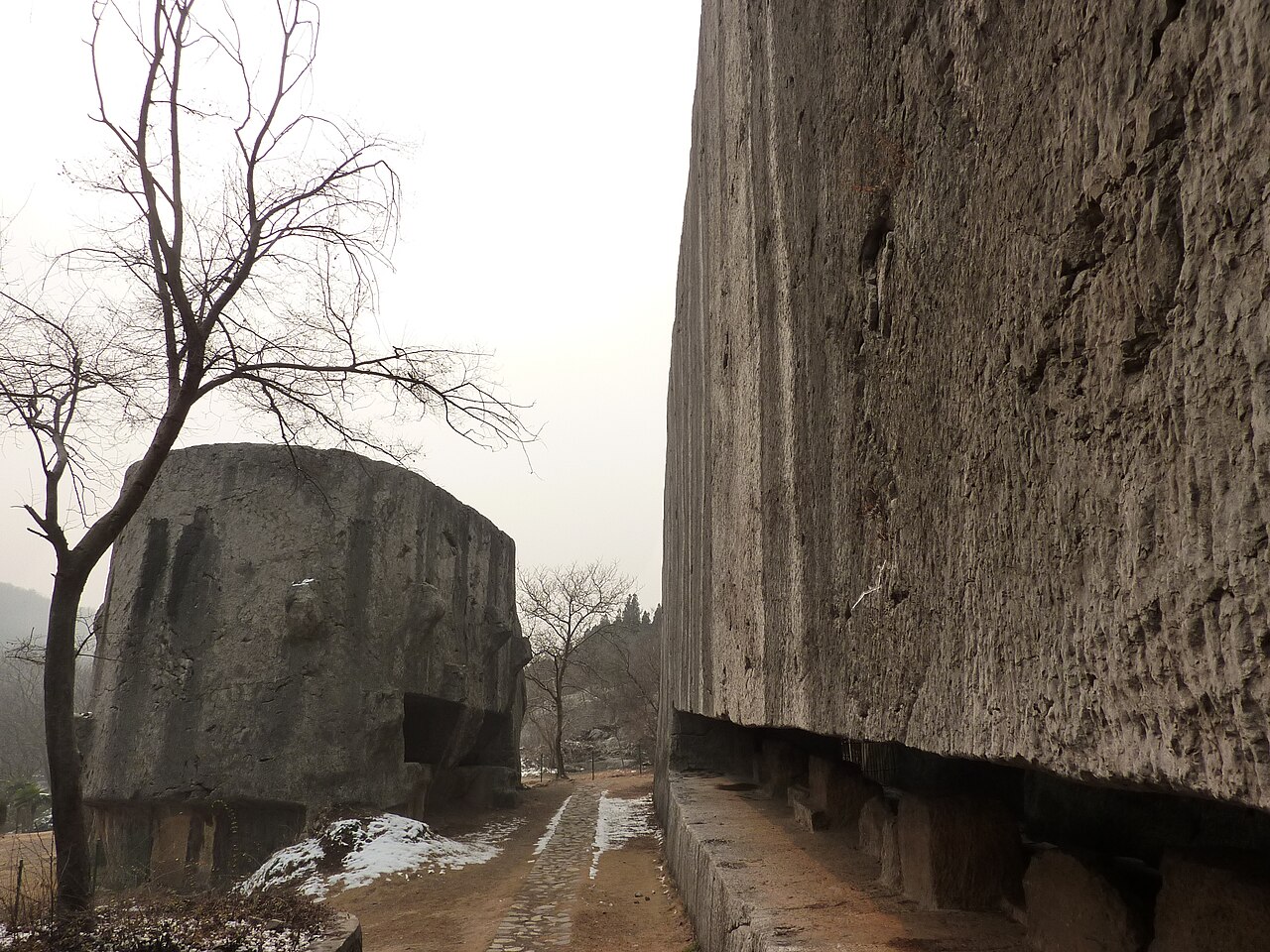 Two colossal cut stone masses at Yangshan Quarry: on the right a towering vertical wall of pale worked rock running away from the camera, undercut at its base and propped on stubs of uncut stone; on the left a huge rounded drum-shaped block standing separately, with a dirt path between them and bare winter trees