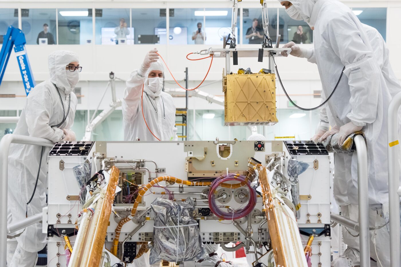 Three engineers in white cleanroom suits and masks guiding a gold box suspended from a crane down into the open white chassis of the Perseverance rover, while colleagues watch from a window above
