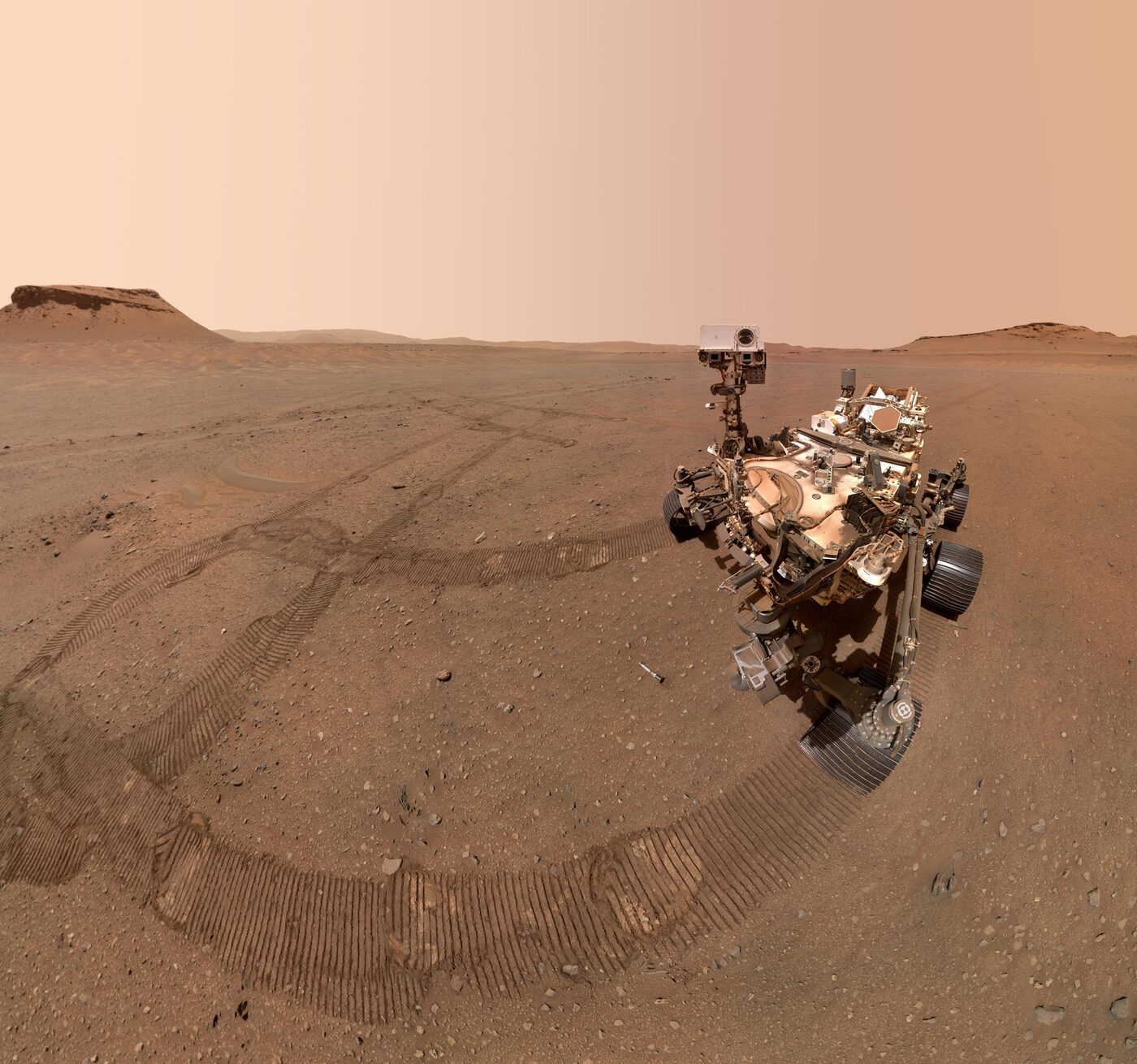 The Perseverance rover photographed from above on the rust-coloured Martian surface, with its wheel tracks curving away across the dust toward low hills on the horizon under a pale peach sky