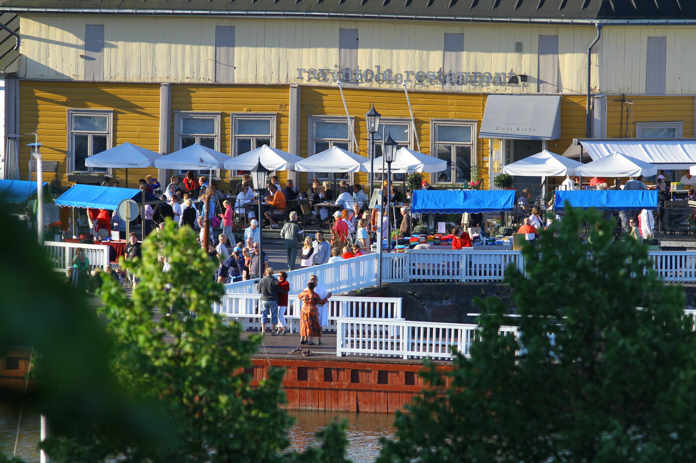 A yellow wooden waterfront building in Naantali Old Town in early morning sunshine, with white café parasols, blue market awnings and a crowd of people standing and sitting along a white-railed boardwalk above the water