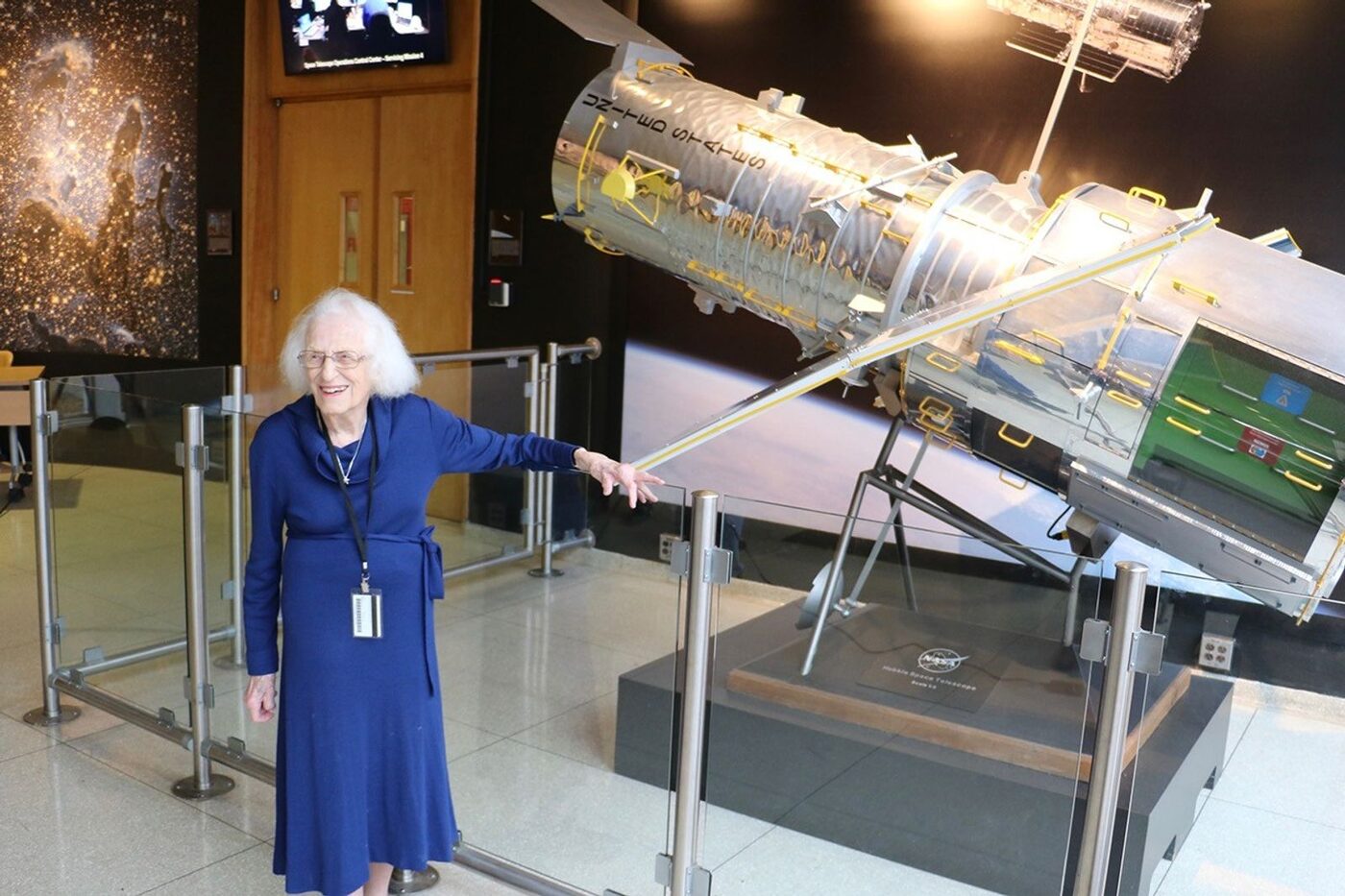 An older woman in a blue dress smiles broadly, one arm raised toward a large silver scale model of the Hubble Space Telescope standing in a building lobby