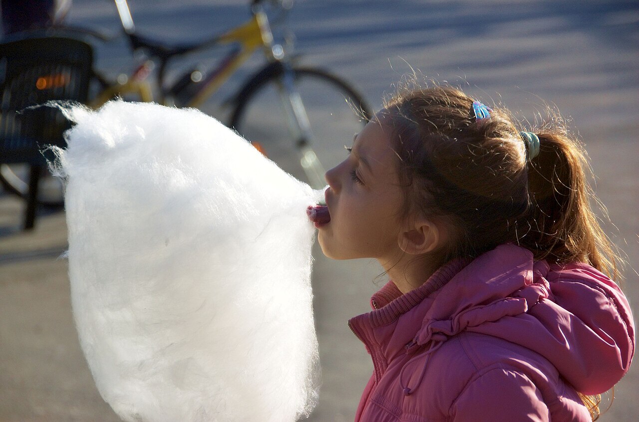 A child at a fair holding an enormous puff of pink cotton candy, mid-bite, beaming