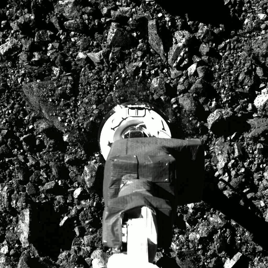 Black and white close-up of a spacecraft's round sampling head at the end of a foil-wrapped robotic arm, hovering just above a surface completely covered in jagged dark rocks and rubble