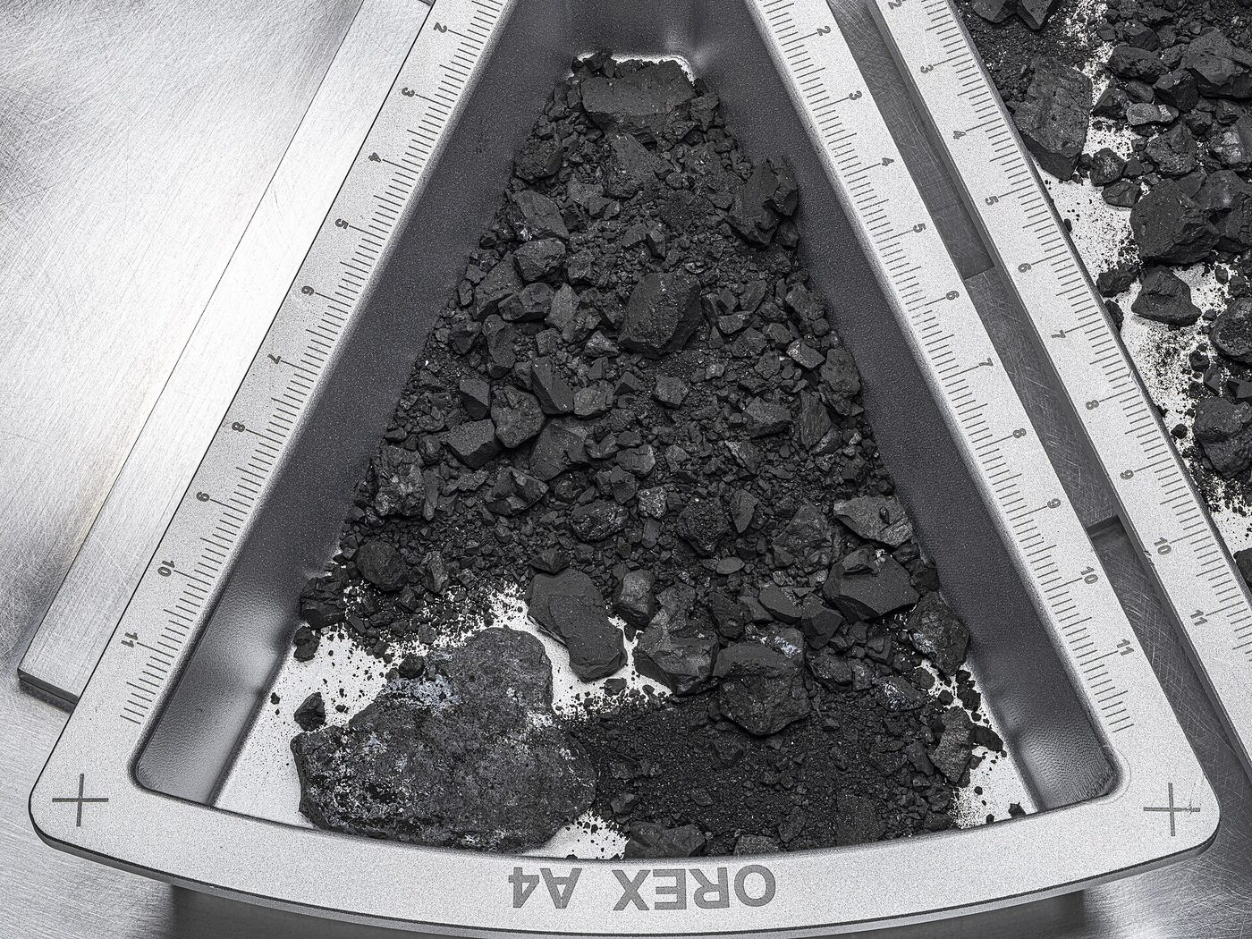 Overhead view of a wedge-shaped metal curation tray filled with jagged coal-black rocks and fine dark dust, with precision rulers laid alongside for scale