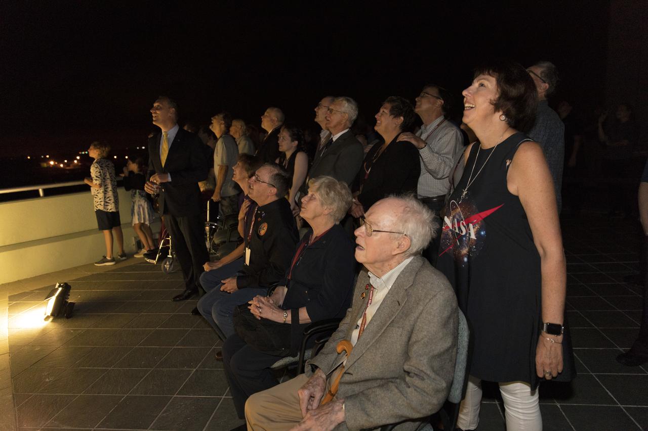 A crowd on a rooftop at night, faces tilted upward and lit by rocket light — Dr. Eugene Parker seated in the foreground, watching the launch of the spacecraft named after him