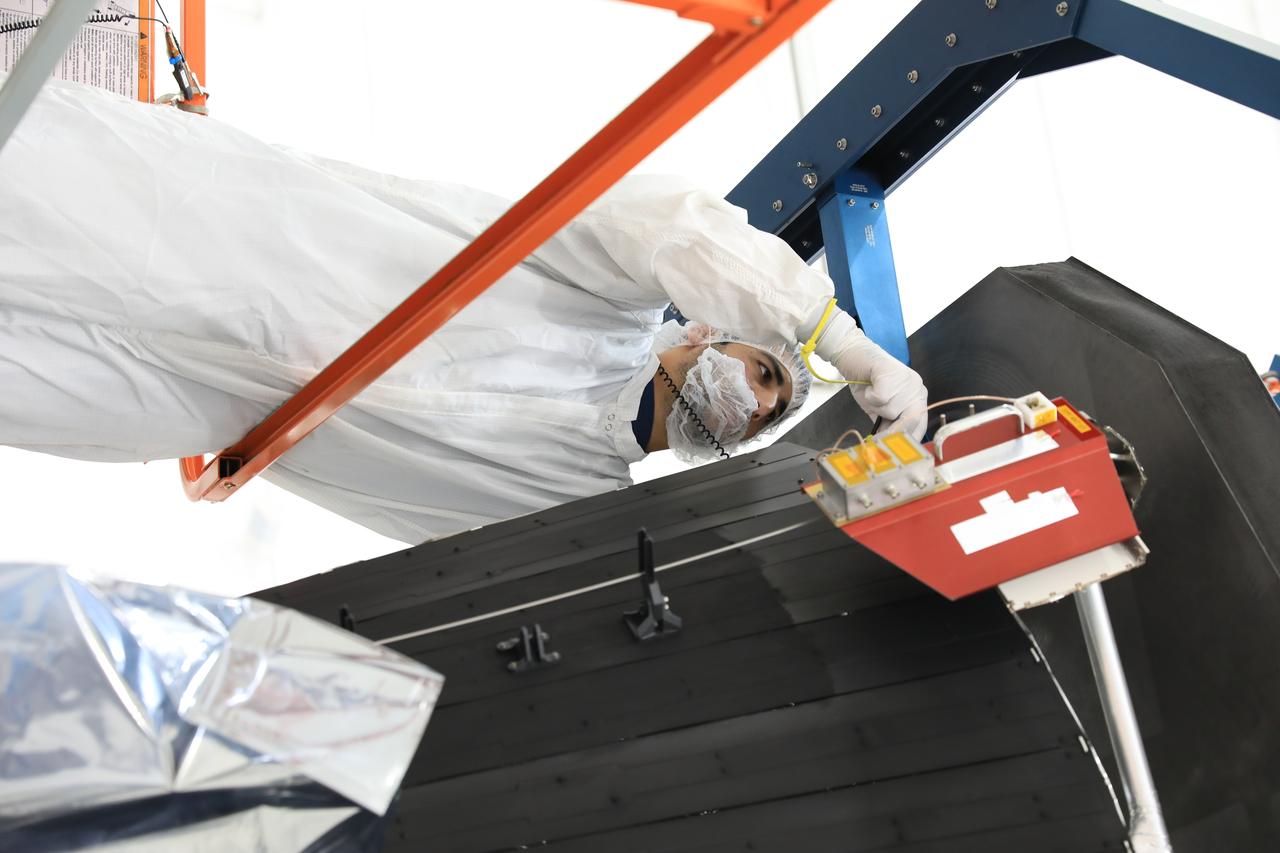 A technician in a white cleanroom suit, hairnet and gloves leaning carefully over the black heat shield of the Parker Solar Probe during installation