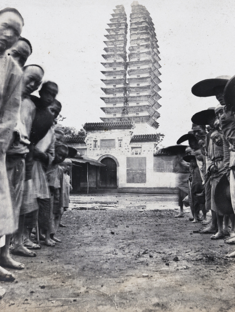 Black and white photograph from the early 1900s: a tall many-tiered brick pagoda split vertically down the middle into two leaning halves, standing behind a temple gateway, with a crowd of people watching from both sides of a bare earth forecourt