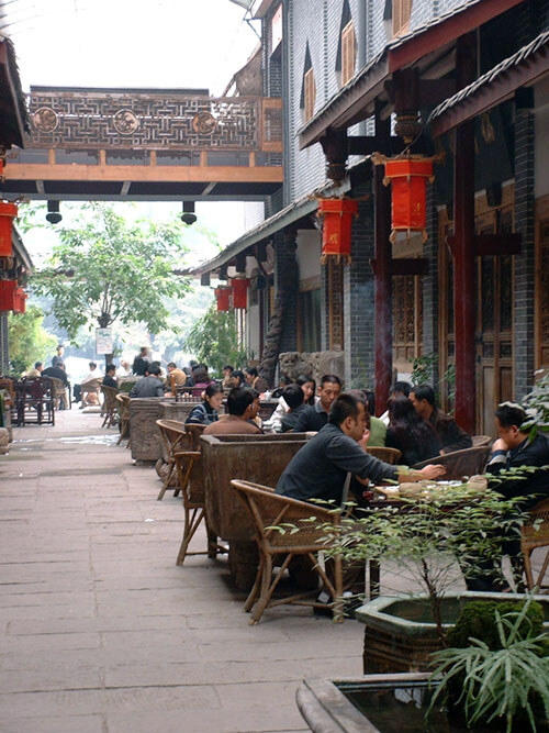 A narrow lane of old timber-fronted shophouses hung with red lanterns, with rows of wicker chairs and low tables along one side where people are sitting drinking tea