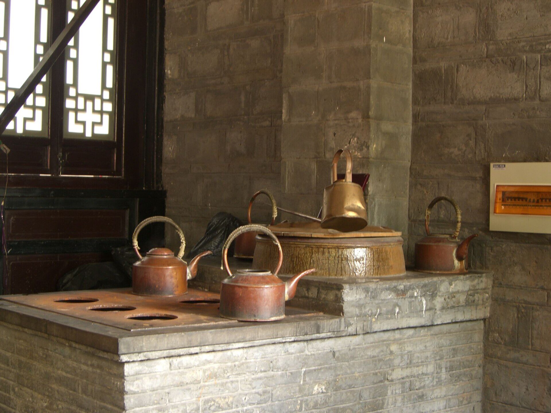 Four copper kettles with tall curved handles sitting on a brick water stove inside a Chengdu teahouse, beside a wooden lattice window