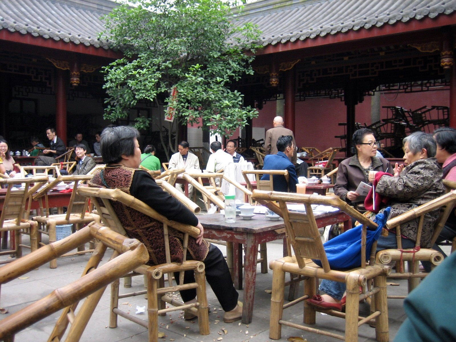 People sitting in bamboo armchairs at wooden tables in the courtyard of a Chinese temple in Chengdu, with red painted timber halls and hanging lanterns behind them