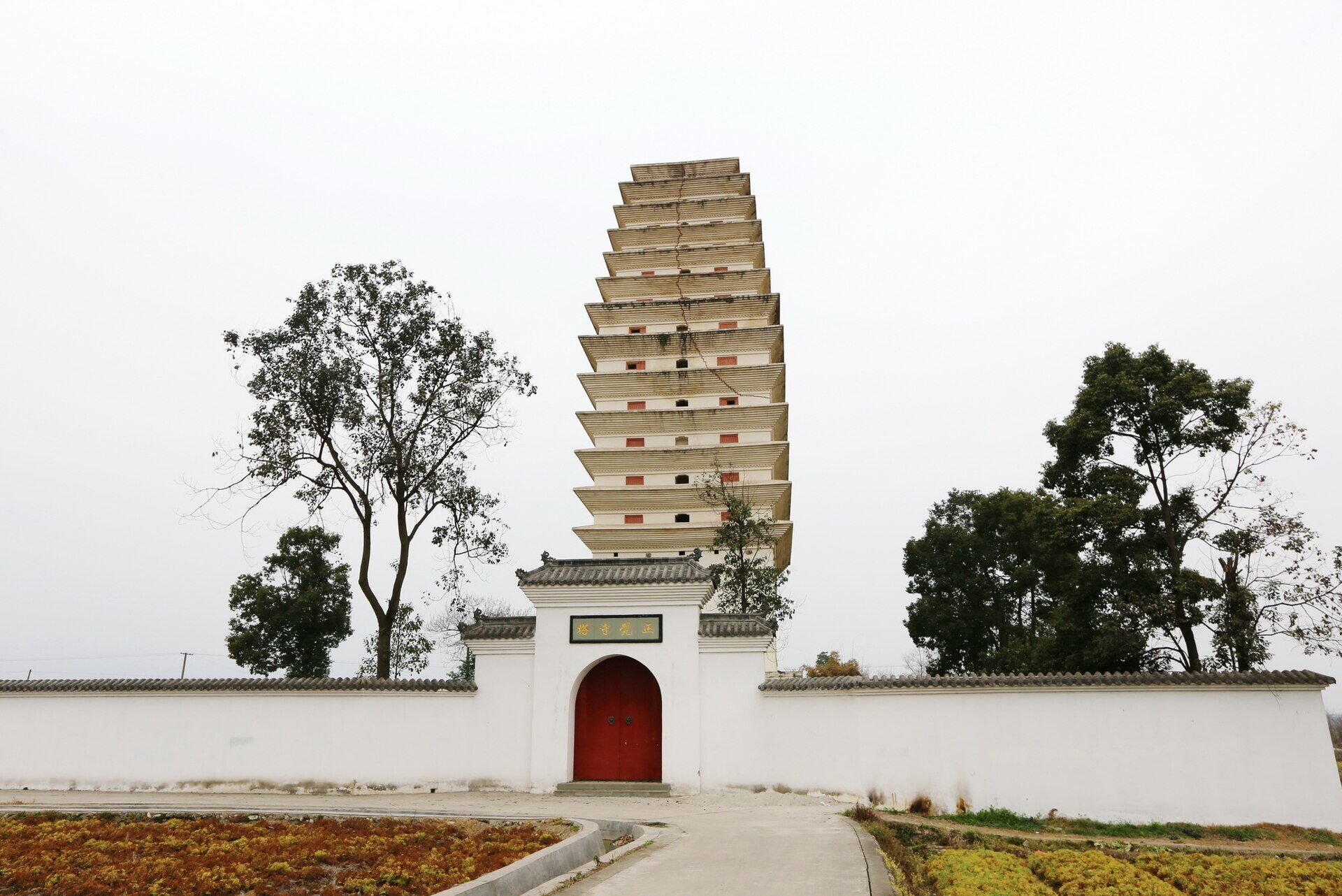 A tall square brick pagoda of many closely spaced tiers rising behind a low white wall with a small red gate, with bare trees to either side and fields in the foreground