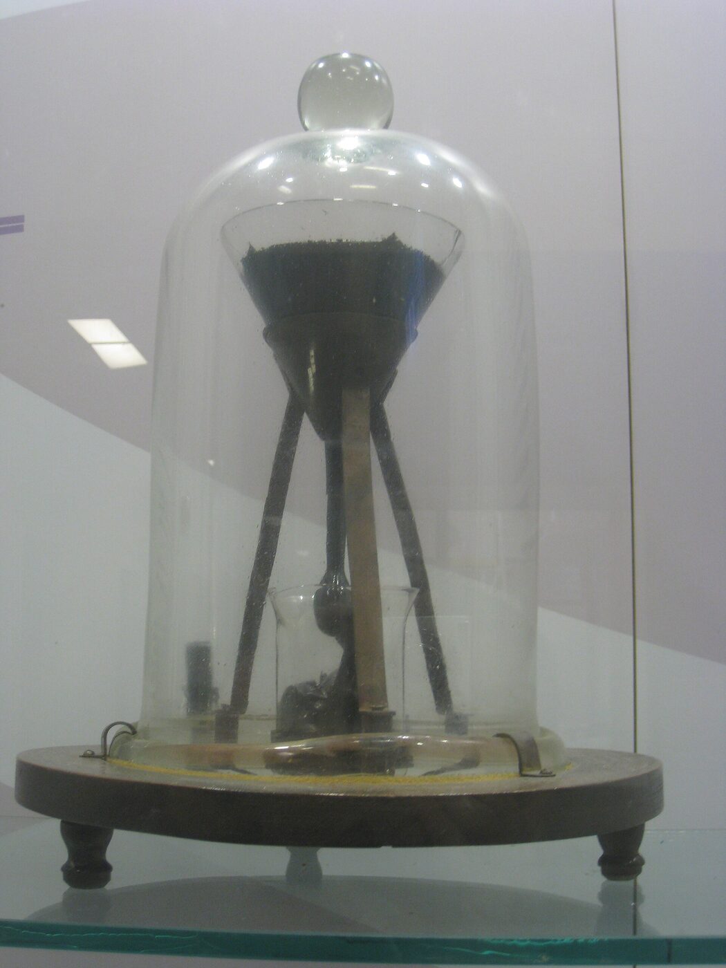 The pitch drop experiment inside its glass display case in 2012, a bell jar over a funnel of black pitch on a wooden stand, with a slender thread of pitch reaching down into a beaker containing a dark pile of earlier drops