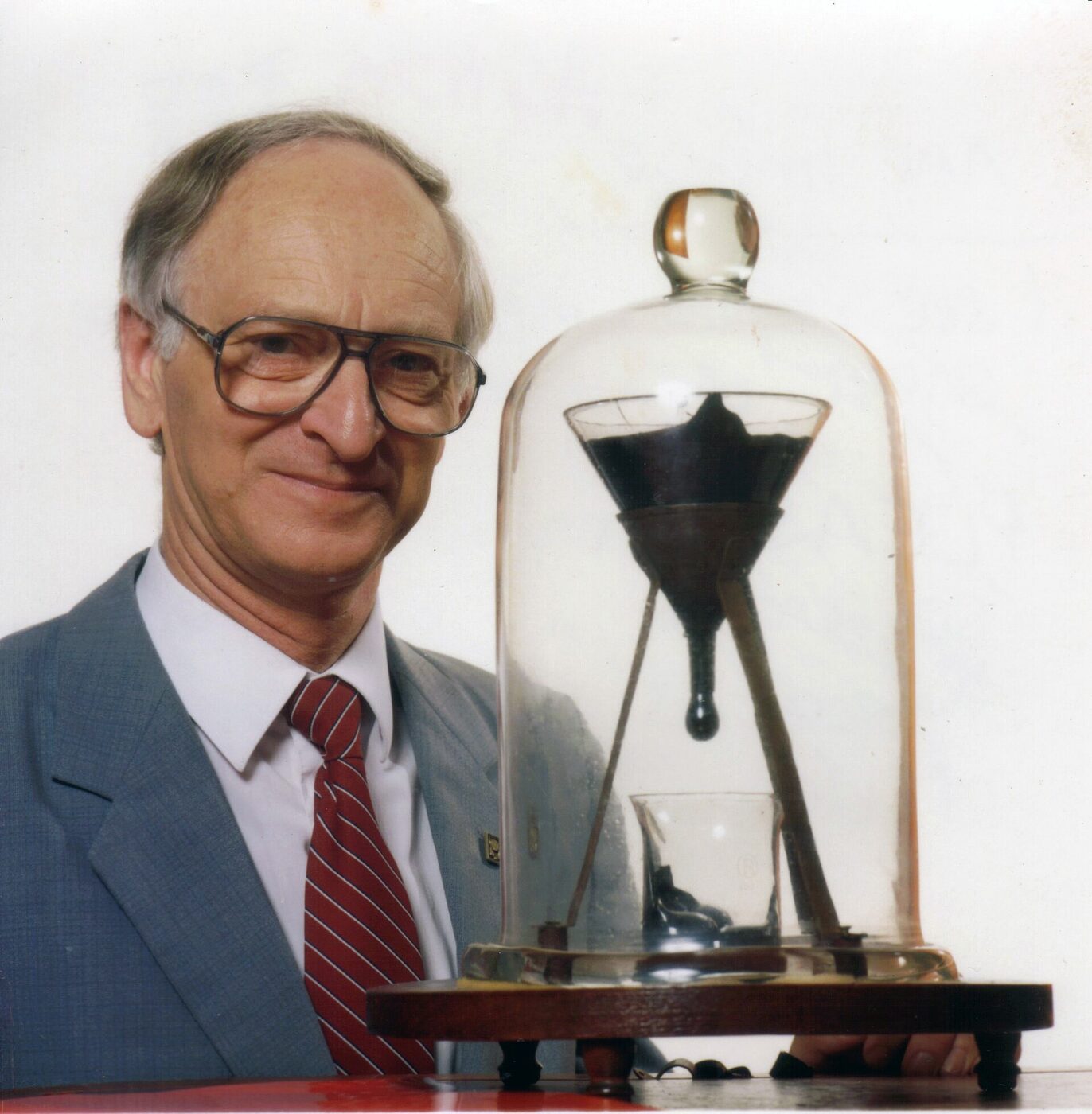 Professor John Mainstone in a grey suit and red striped tie, smiling gently beside the glass bell jar containing the pitch drop apparatus, with a small rounded bead of black pitch hanging from the funnel stem