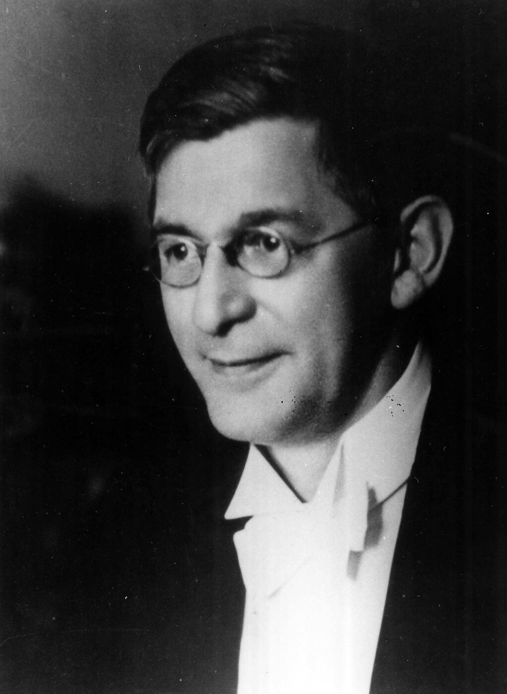 Black and white portrait photograph of Thomas Parnell around 1920, a young man in a dark suit and white collar wearing round wire-rimmed glasses, turned slightly to one side with a faint amused smile