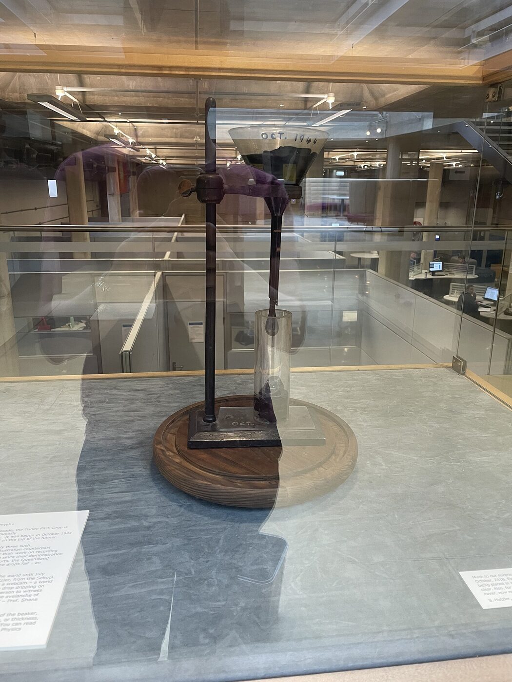 The Trinity College Dublin pitch drop experiment in a glass display case in a library, a funnel labelled OCT. 1944 in handwriting, clamped to a stand with a long dark rod of pitch descending into a glass cylinder below