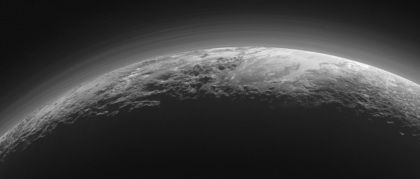 Near-sunset view across Pluto's horizon — jagged icy mountains rising above smooth frozen plains, with layers of blue haze in the sky