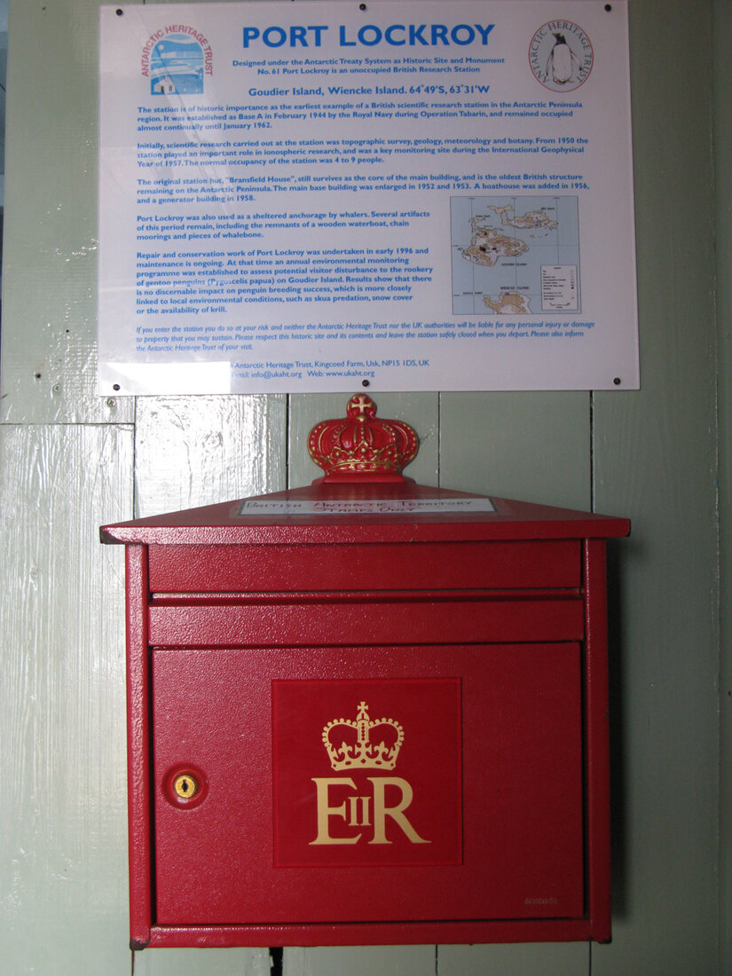 A red British wall-mounted post box with a gold EIIR crown insignia, fixed to a pale wooden wall beneath a printed Port Lockroy information notice