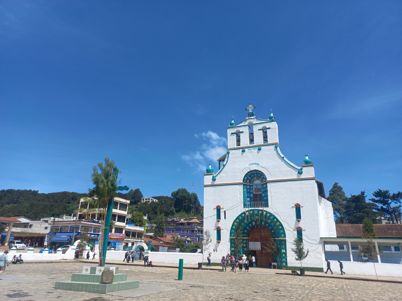 The whitewashed church of San Juan Bautista in San Juan Chamula, its façade painted with turquoise and green floral patterns and dated 1522 1524, under a bright blue sky with the town plaza in front
