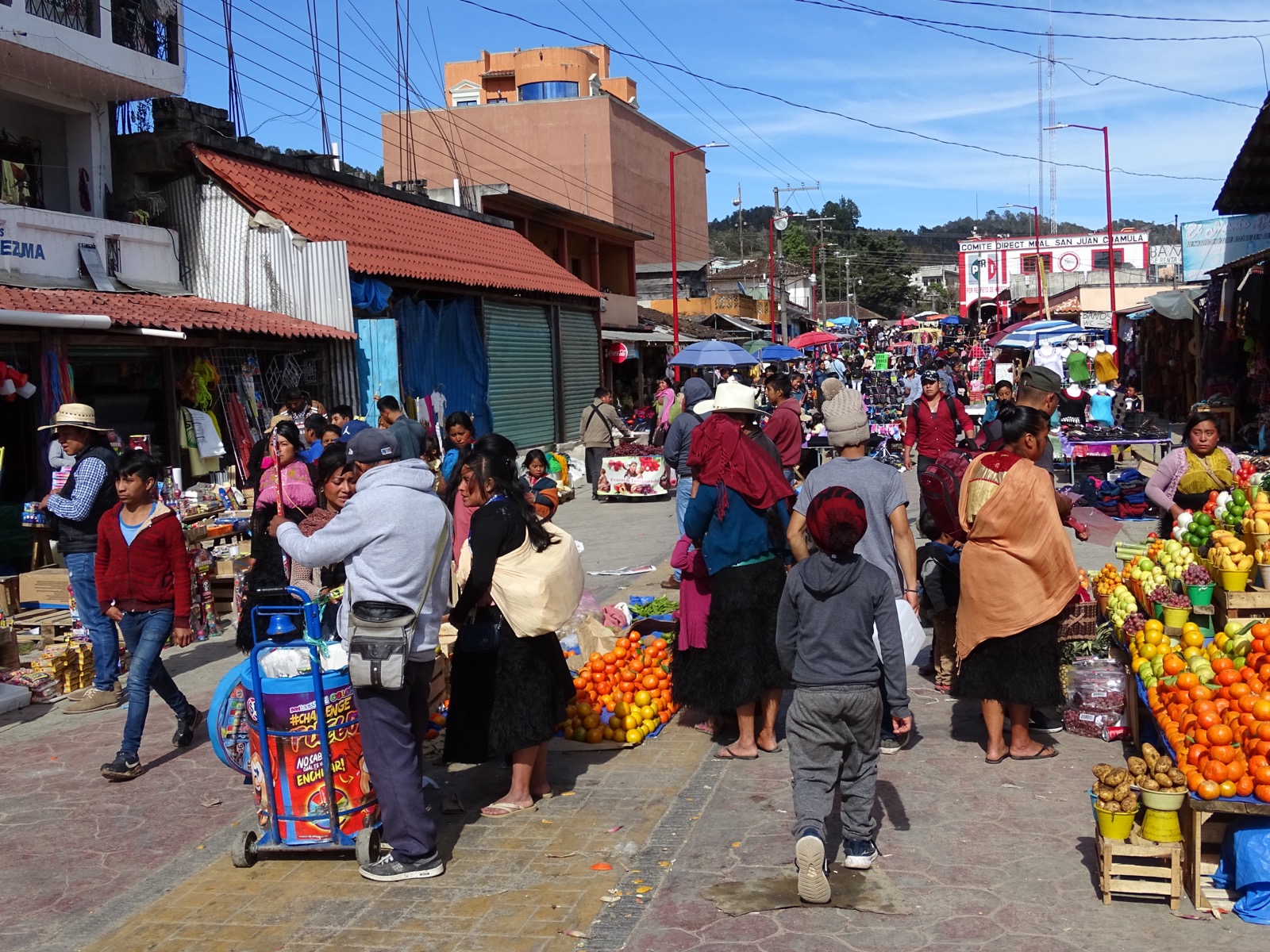 A busy market street in San Juan Chamula crowded with people, fruit stalls of oranges and limes, umbrellas and a sign reading Comité Directivo Municipal San Juan Chamula