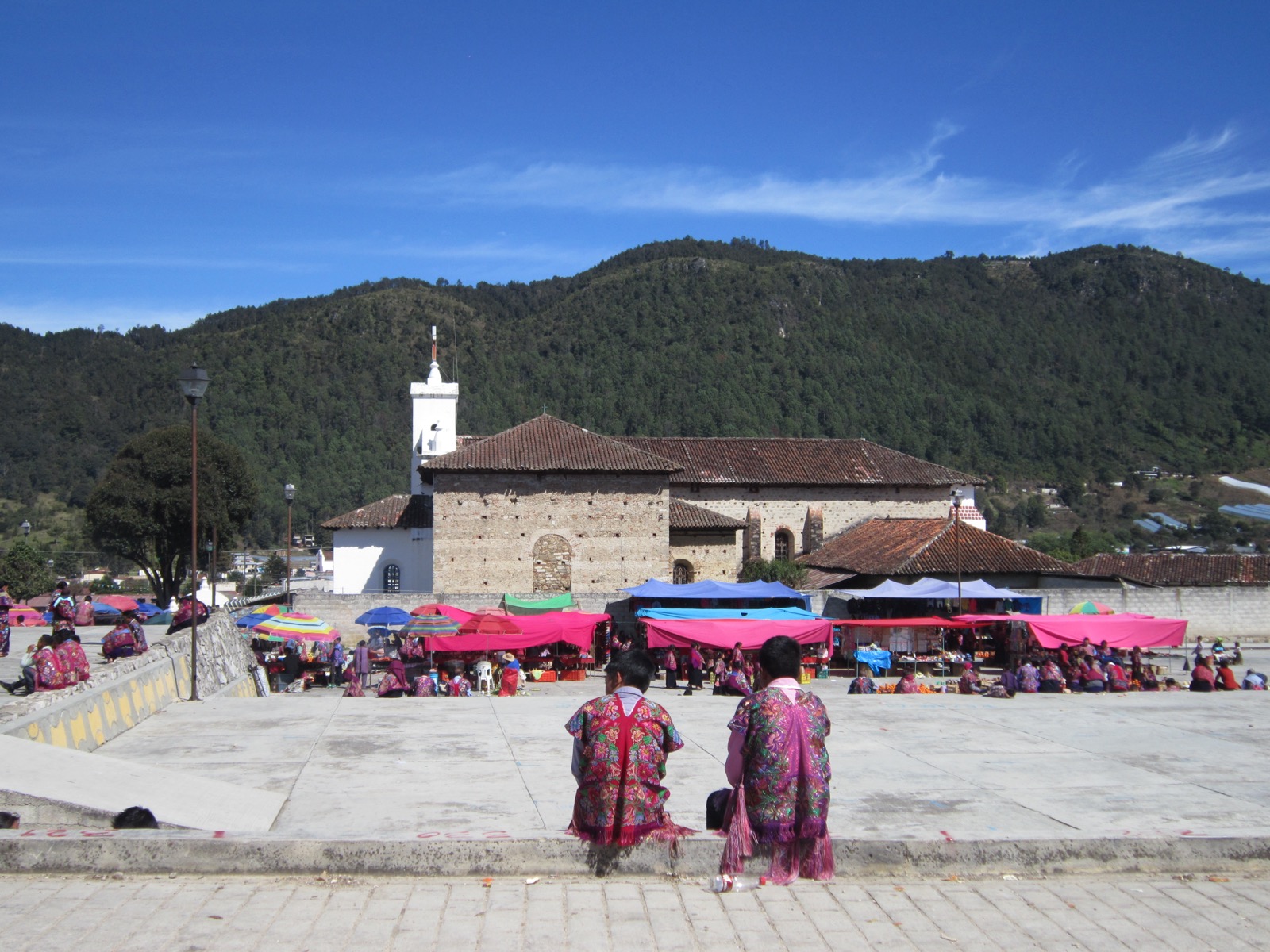 The stone church of Zinacantán with forested mountains behind it and a market of pink and blue awnings in the plaza, two people in bright floral embroidered capes seated in the foreground