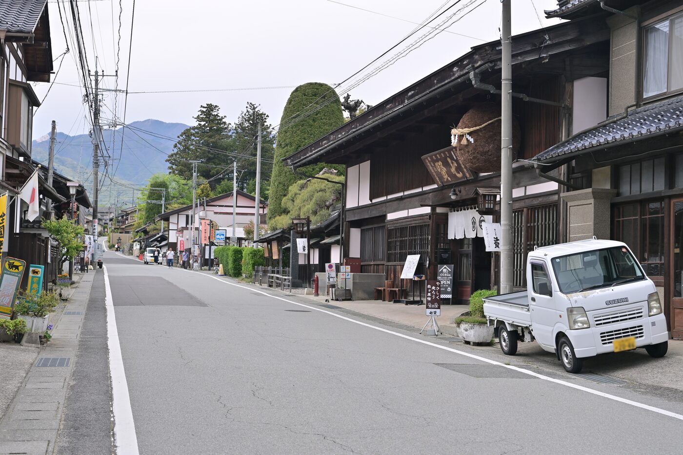 A quiet two-lane street through an old Japanese post town, lined with dark-timbered traditional shopfronts and noren curtains, with green mountains rising in the haze at the end of the road