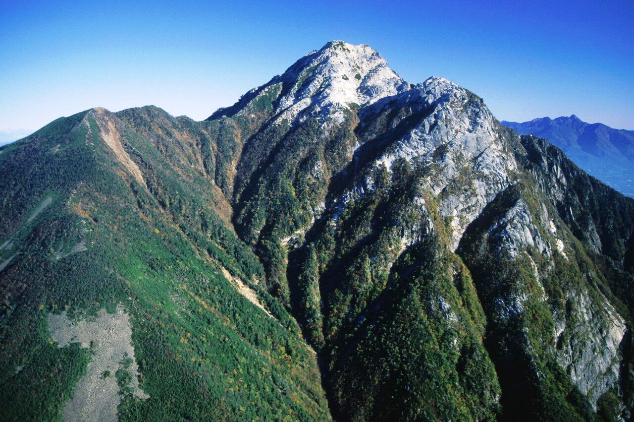 A steep granite peak with pale white rock near its summit rising above dark green forested ridges, under a deep blue sky