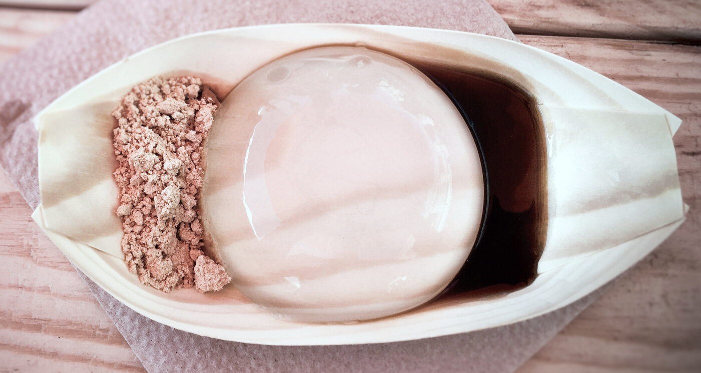 Overhead view of a clear rounded water jelly in a small boat-shaped wooden dish, with a heap of pale roasted soybean flour on one side and a dark pool of black sugar syrup on the other