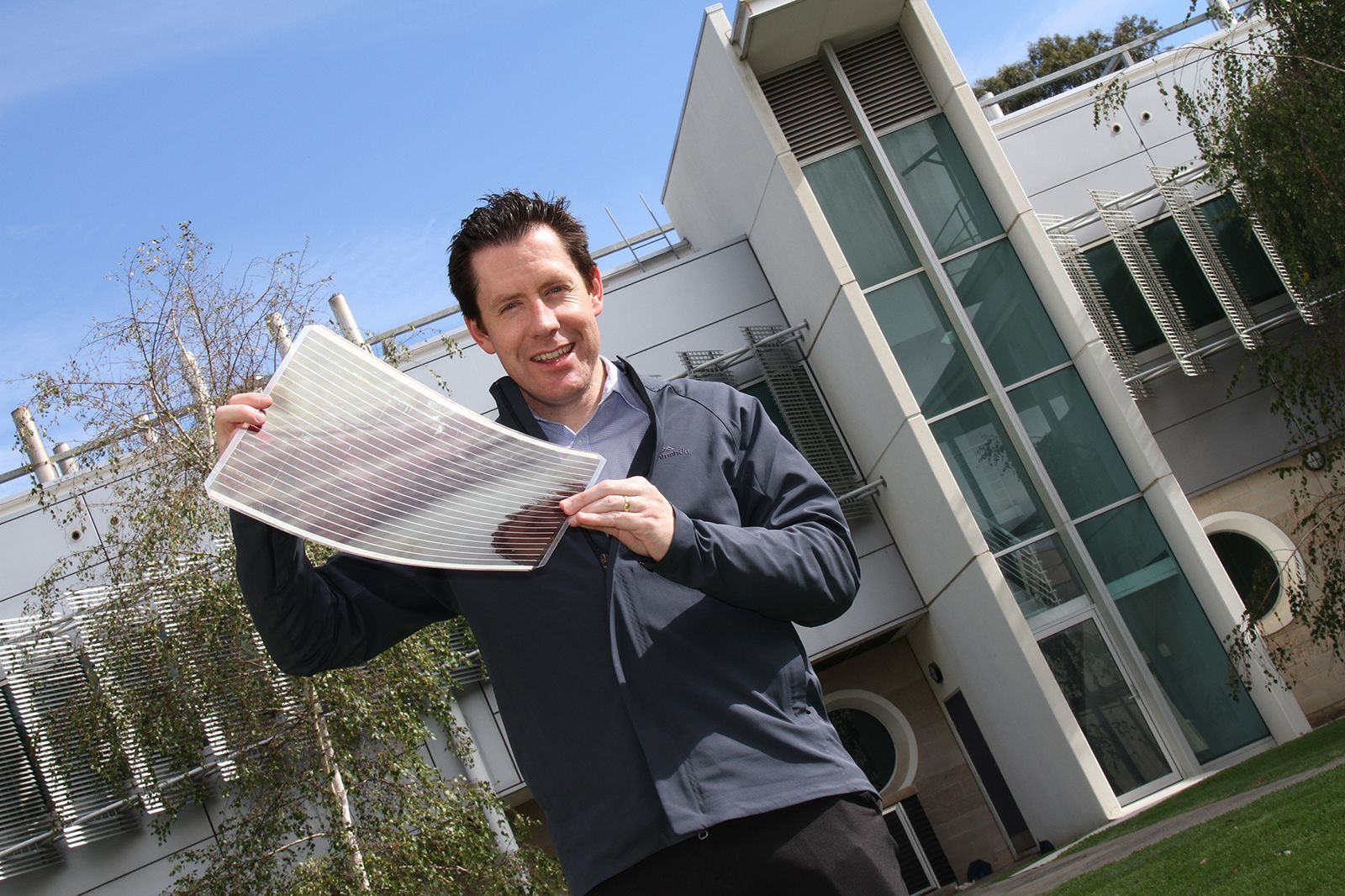 A researcher outdoors holding up a large flexible sheet of printed plastic solar cells, showing rows of dark stripes on a clear film