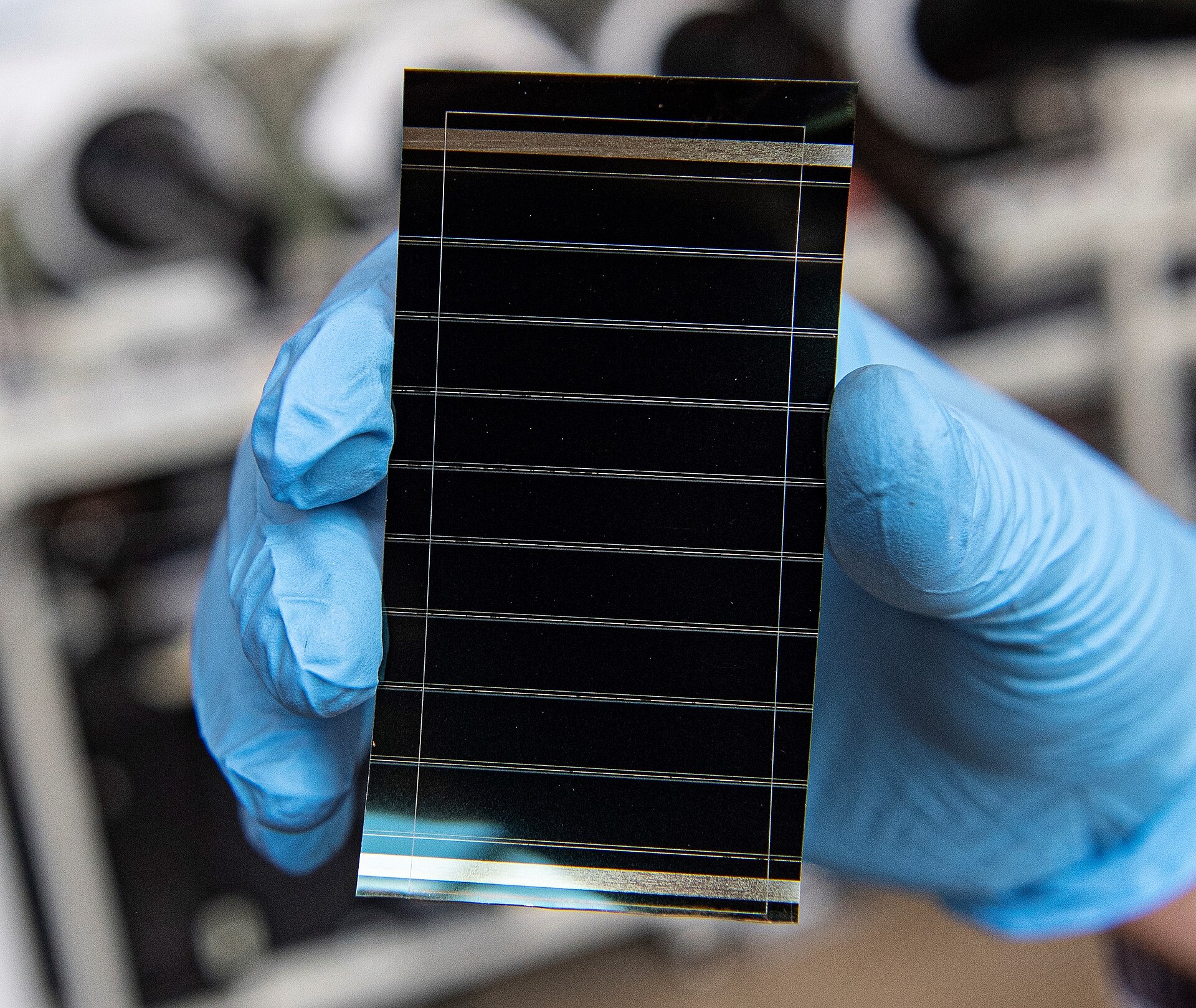 Two blue-gloved hands holding a small rectangular perovskite solar cell on glass, its dark surface crossed by fine parallel silver lines