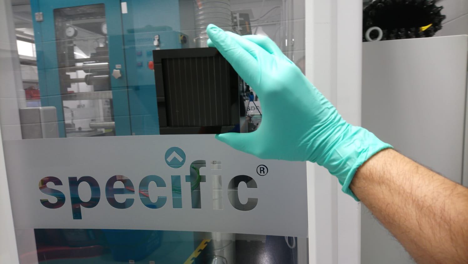 A green-gloved hand holding a small square dark perovskite solar module, roughly ten centimetres across, in a laboratory with equipment behind