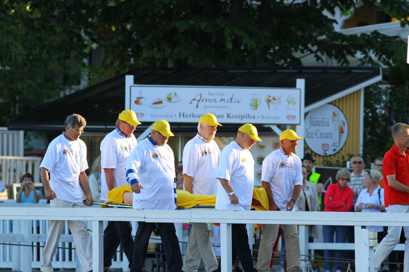 Six older men in matching white shirts and bright yellow baseball caps walking in step along a white-railed waterfront boardwalk, carrying a stretcher between them; a yellow blanket covers the person on it and one bare leg is visible
