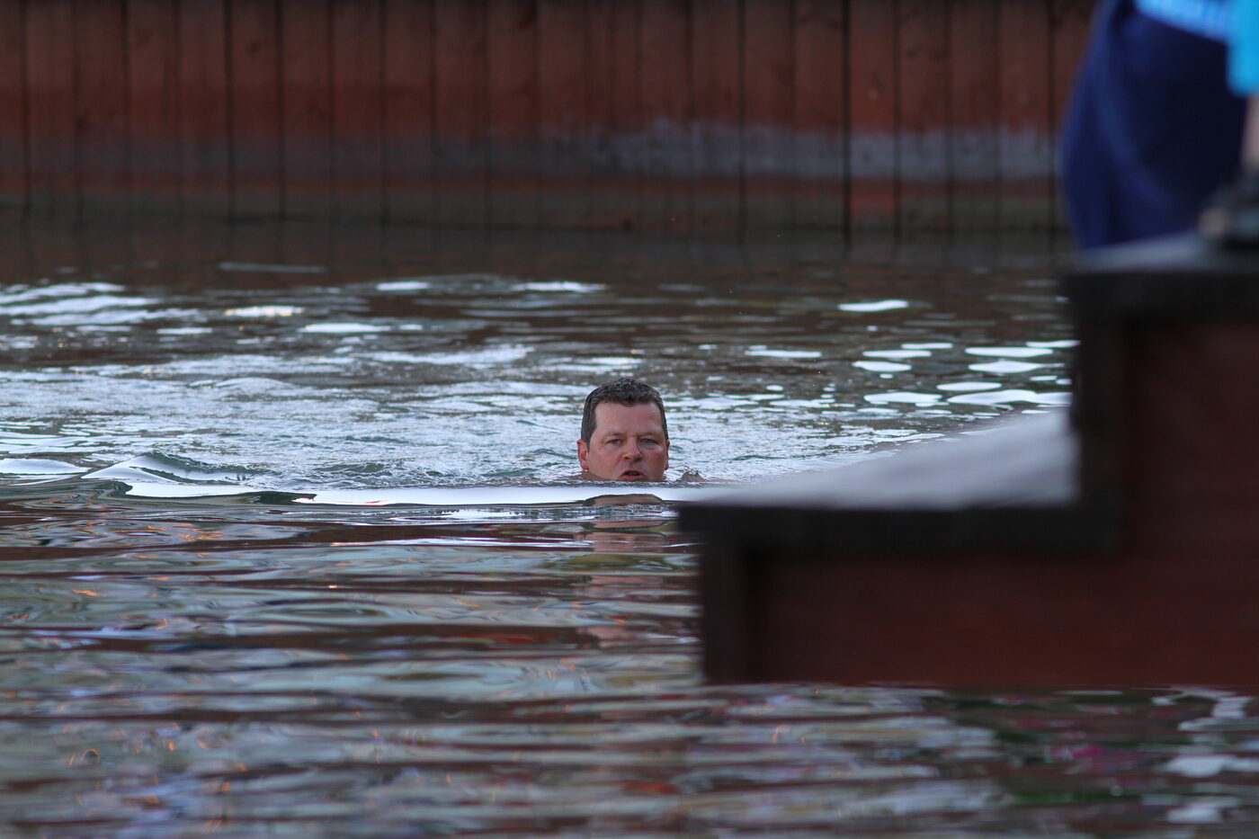 A man's head and shoulders just above the surface of calm harbour water, swimming back toward a wooden quay and a set of steps at the right of frame