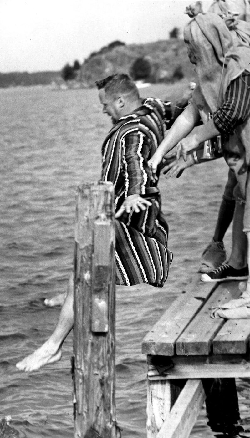 Black and white photograph from 1967 of a bearded man in a striped dressing gown being tipped off the end of a wooden jetty into the sea, barefoot, with several pairs of hands reaching from the right to help him on his way