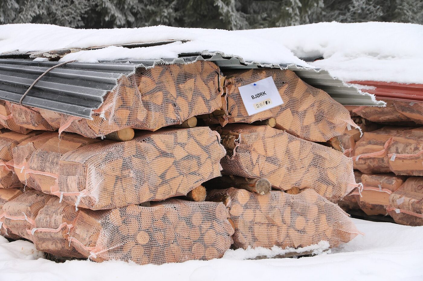 Stacked mesh sacks of split Norwegian birch firewood under a corrugated metal sheet in deep snow, with a handwritten paper label reading BJØRK tucked into one of the sacks