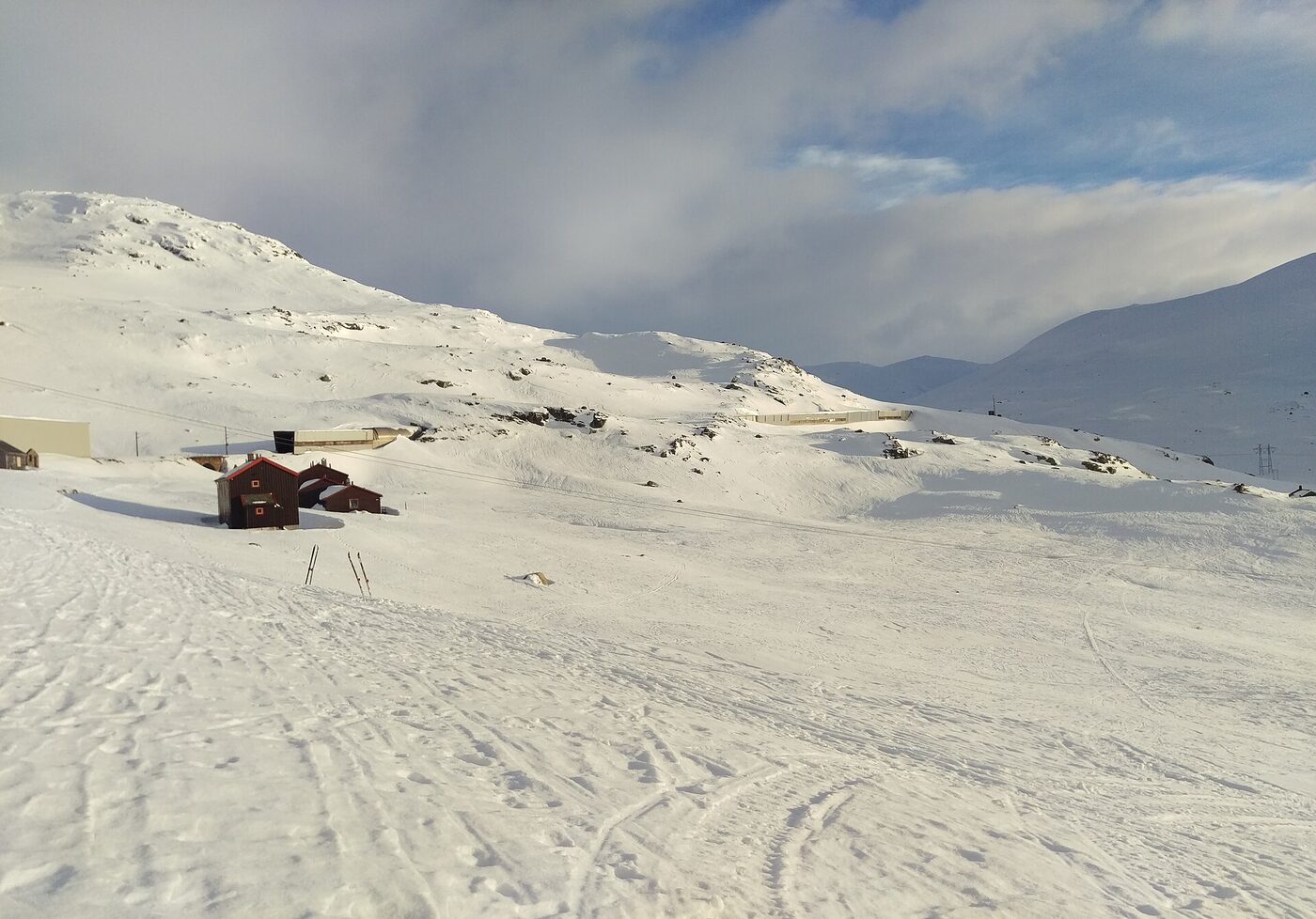 A snow-covered high plateau in bright winter light, with a small dark-red timber cabin half-buried in drifts, a pair of cross-country skis planted upright in the snow, and the long low concrete snow shelter of the Bergen Line running along the ridge behind