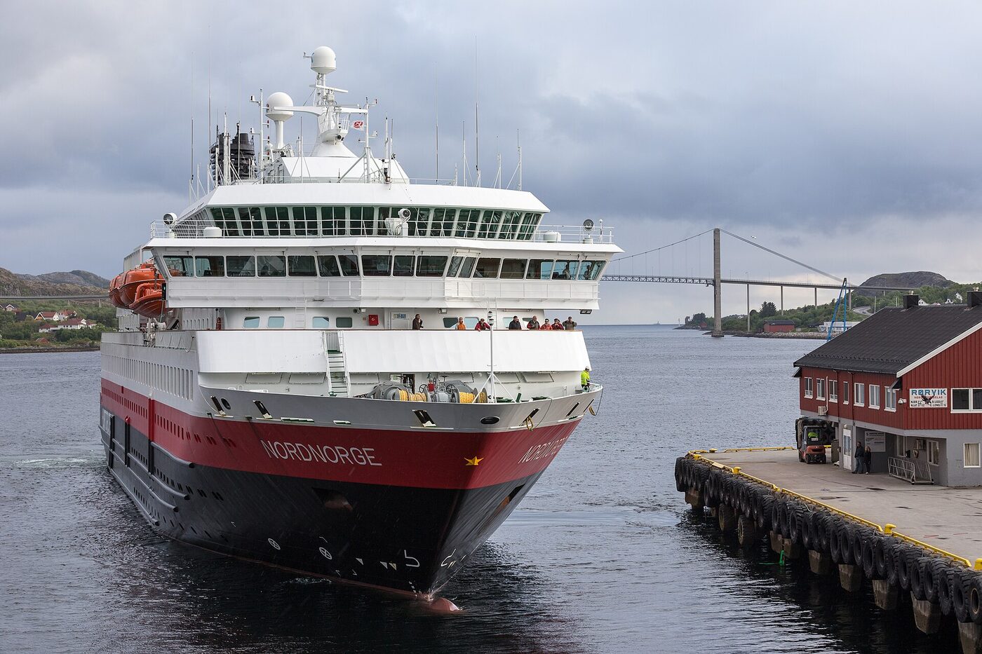 MS Nordnorge coming bow-first alongside a concrete quay at Rørvik, its red-and-black hull filling the frame, crew in high-visibility gear on the foredeck, passengers watching from the open deck below the bridge, and a suspension bridge across the sound behind