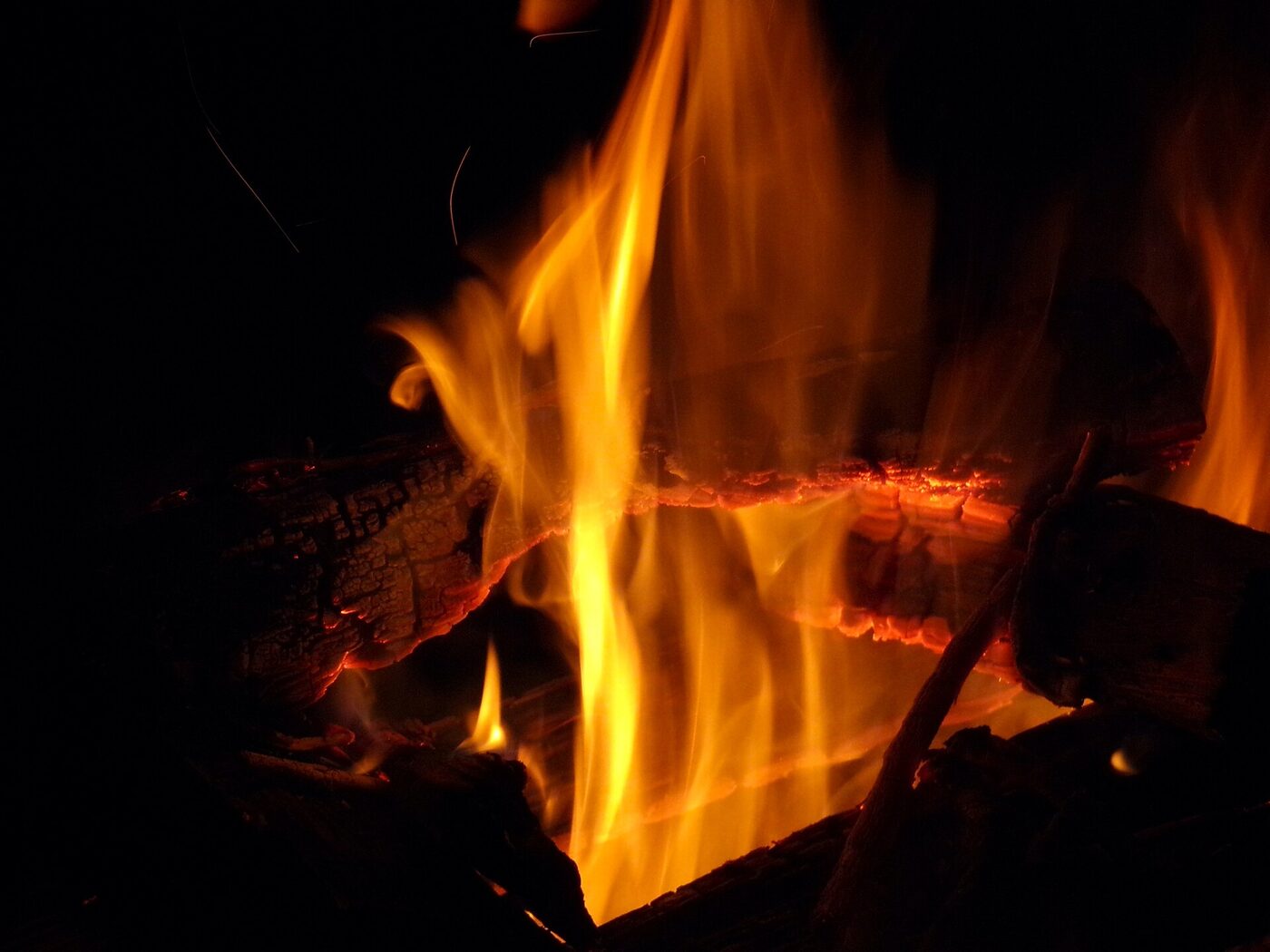 Close-up of a wood fire at night, tall yellow flames rising between two split logs whose edges glow orange along the cracks, everything else in darkness