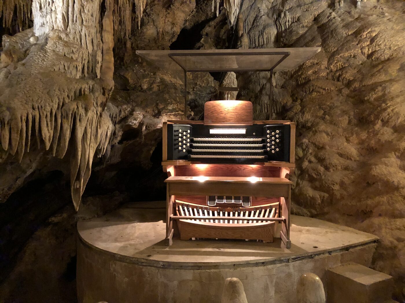 Wide view of the Great Stalacpipe Organ console lit on a concrete platform, with a canopy overhead and thick draperies of stalactites hanging on the cave wall to the left