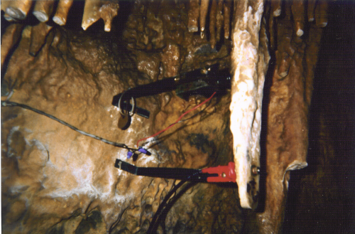 Close-up of a black solenoid striker with red and black wires, bolted to the cave rock beside a pale stalactite