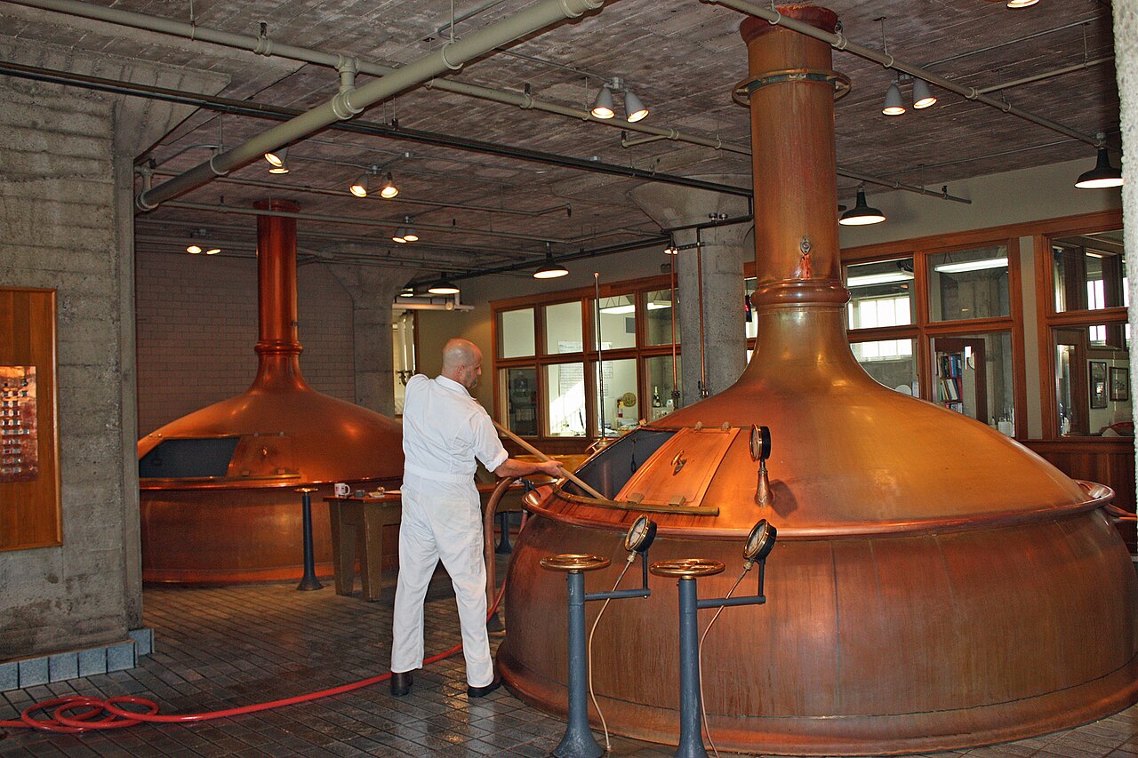 A brewer in white overalls leaning over a large polished copper brewing kettle, with a second copper kettle behind and a brick ceiling above