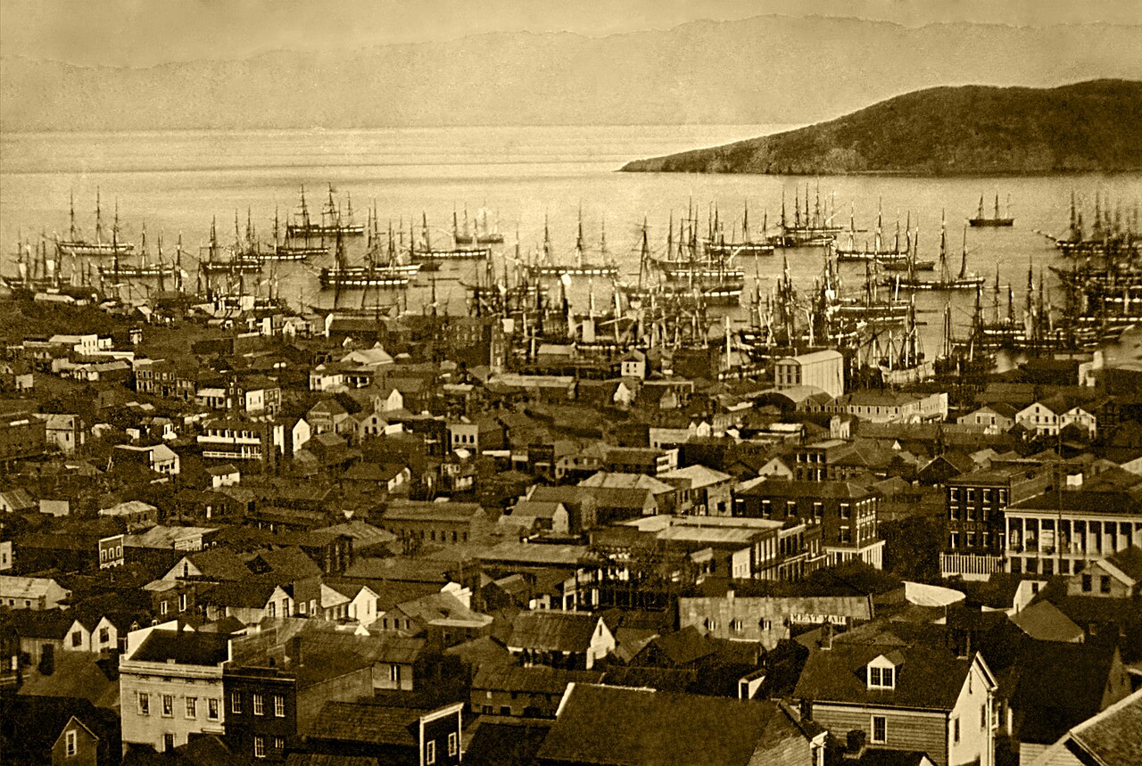 Sepia daguerreotype looking down over the rooftops of 1850s San Francisco toward a bay crowded with the masts of tall ships