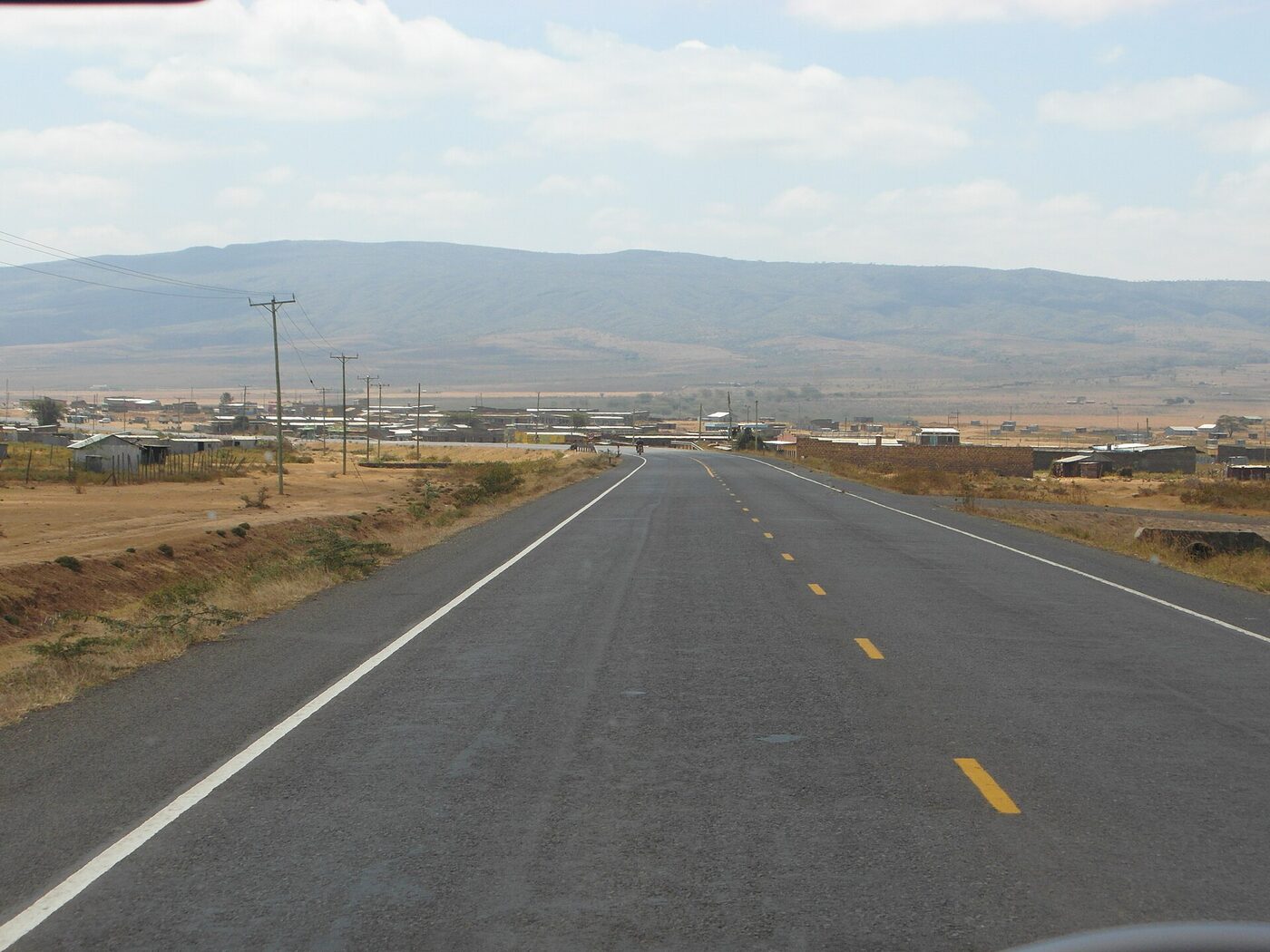 A straight two-lane tarmac road running through dry scrubland near Narok, Kenya, with a small roadside settlement of tin-roofed buildings and hazy hills on the horizon
