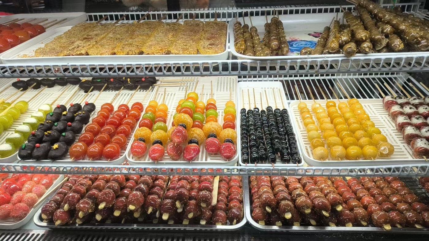 A refrigerated display case packed with trays of candied fruit skewers — strawberries, grapes, oranges, blueberries and chocolate-dipped varieties in many colours