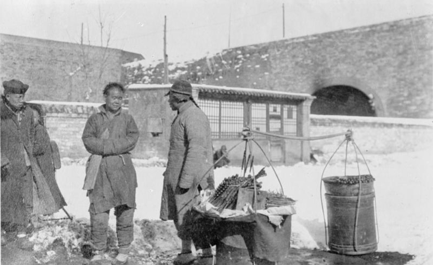 Black-and-white photograph from around 1900 of a street vendor in Peking standing in the snow with a carrying pole and bundles of candied fruit skewers