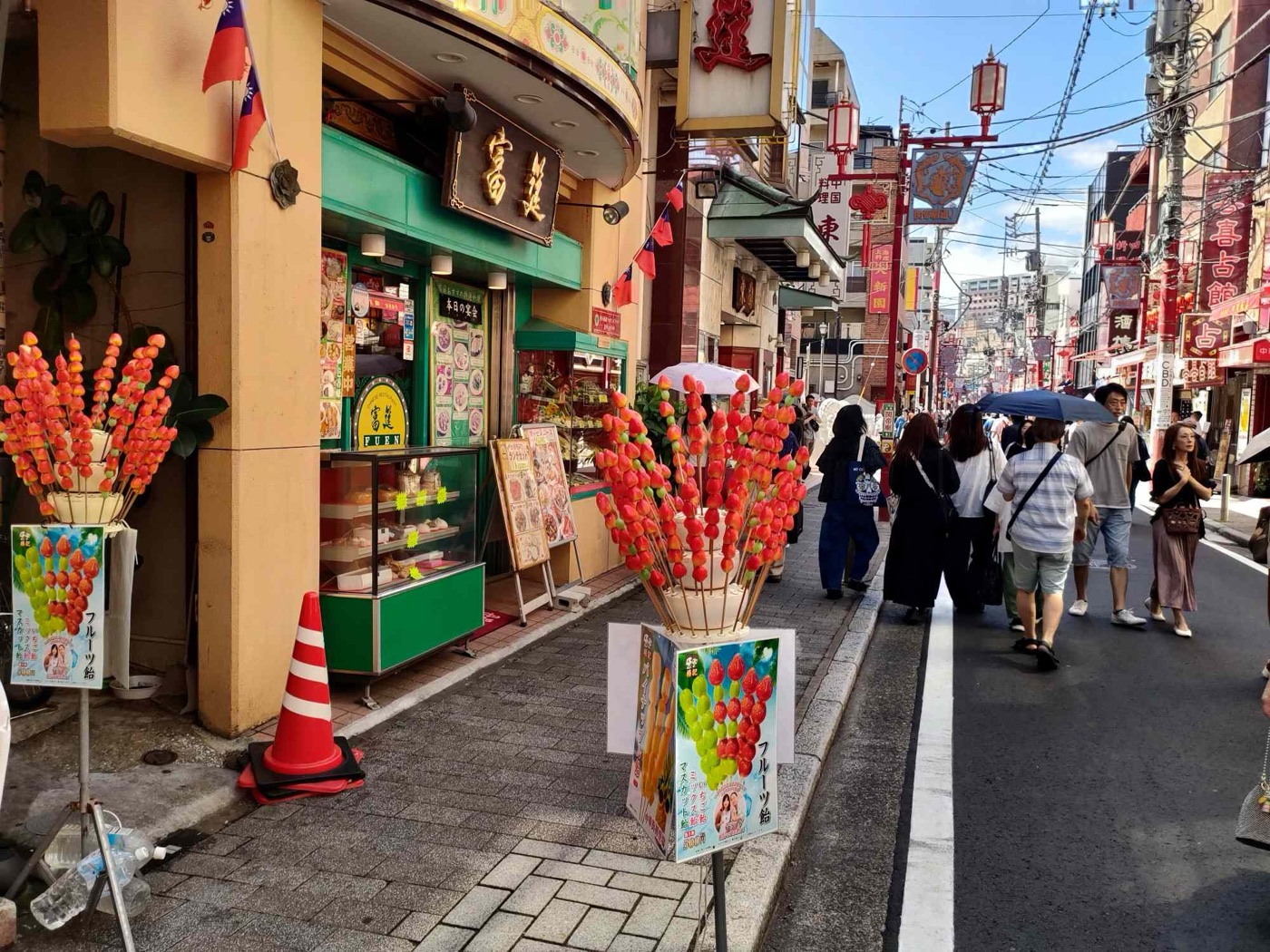 A busy Chinatown street in Yokohama, Japan, with two tall bouquets of red candied strawberry skewers displayed on the pavement outside a shop